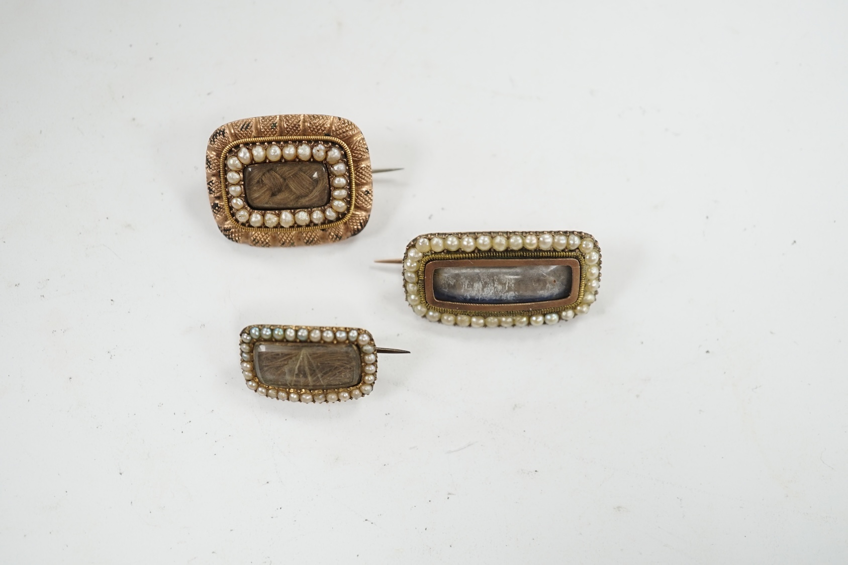 Three assorted 19th century yellow metal and seed pearl set mourning brooches, two with plaited hair beneath a glazed panel, largest 27mm. Condition - poor to fair